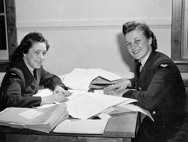 Black and white photograph. Two women, wearing RCAF uniforms but no caps, sit across from each other at a table covered in piles of paperwork.